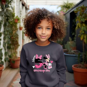 May include: A dark gray sweatshirt with a pink and white Minnie Mouse design. The design features Minnie Mouse holding a pink gift bag and a pink heart balloon. The number 5 is in the design and the text "Name Birthday Girl" is below the design.