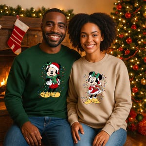 K&ouml;nnte beinhalten: Zwei Personen tragen Sweatshirts zum Thema Weihnachten. Eines ist dunkelgr&uuml;n, das andere ist beige. Beide zeigen einen Cartoon von Mickey und Minnie Mouse in Weihnachtsoutfits, umgeben von Lichterketten. Im Hintergrund ist ein Weihnachtsbaum zu sehen.