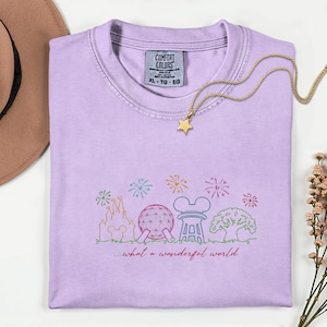 May include: A light purple Comfort Colors t-shirt with a colorful embroidered design of a castle, Spaceship Earth, and other theme park icons. The text "...what a wonderful world" is below the design. A gold star pendant necklace rests on the shirt.