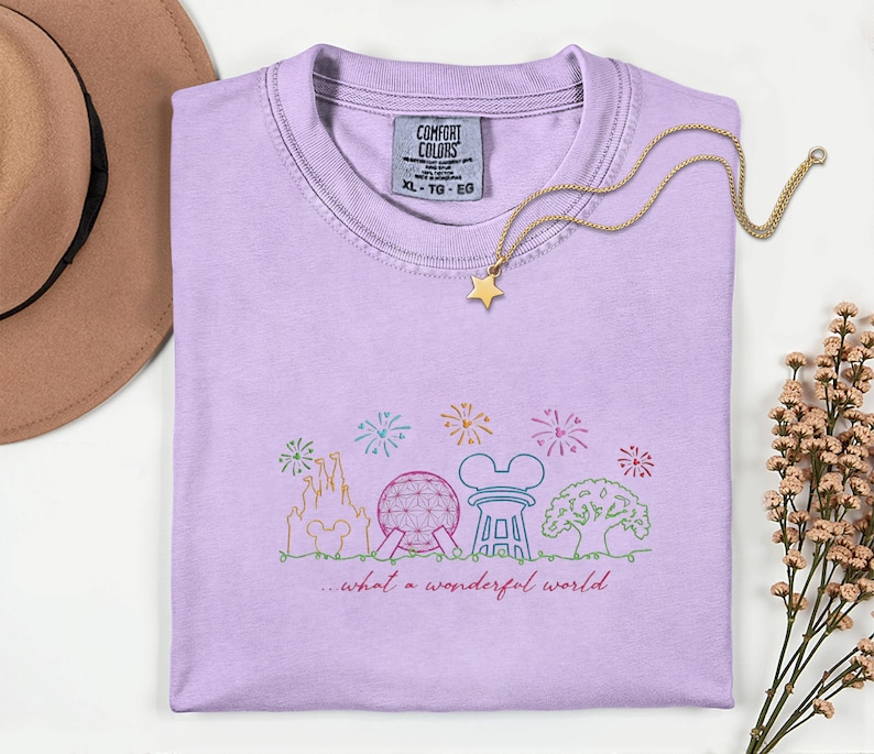 May include: A light purple Comfort Colors t-shirt with a colorful embroidered design of a castle, Spaceship Earth, and other theme park icons. The text "...what a wonderful world" is below the design. A gold star pendant necklace rests on the shirt.