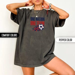 May include: A dark gray t-shirt with the text "ONE NATION ONE TEAM" in red and blue. A soccer ball with red and blue streaks is below the text. The shirt is labeled "Comfort Colors" and "Pepper Color".