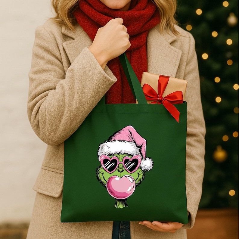 May include: A dark green tote bag featuring a cartoon Grinch character wearing a pink Santa hat and heart-shaped sunglasses, blowing a pink bubble. A small gift wrapped in brown paper with a red bow is inside the bag.