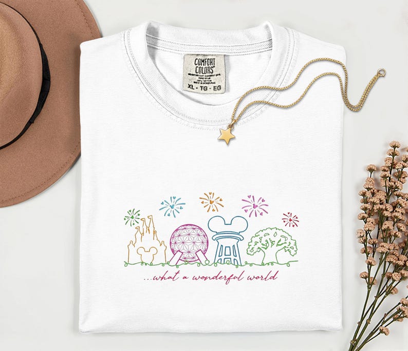 May include: White Comfort Colors t-shirt with a colorful line drawing of a castle, sphere, and other theme park icons. The text "...what a wonderful world" is below the drawing. A gold star necklace is on the shirt.