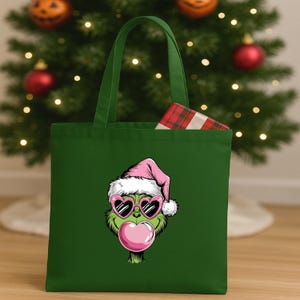 May include: A green tote bag featuring a cartoon Grinch face wearing heart-shaped sunglasses and a pink Santa hat, blowing a pink bubble. A wrapped gift is inside the bag. The bag is in front of a Christmas tree.