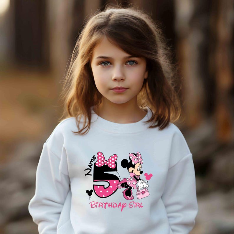May include: White sweatshirt with a pink and black Minnie Mouse graphic. The graphic features Minnie Mouse holding a pink purse and a pink bow. The text "Name" and "Birthday Girl" are printed on the sweatshirt.