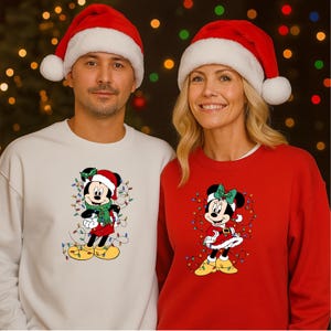 K&ouml;nnte beinhalten: Passende Weihnachts-Sweatshirts mit Mickey und Minnie Mouse. Die Sweatshirts sind cremefarben und rot, wobei Mickey einen gr&uuml;nen Schal und Minnie eine gr&uuml;ne Schleife tr&auml;gt. Beide Charaktere tragen Weihnachtsm&uuml;tzen und sind von Lichterketten umgeben.