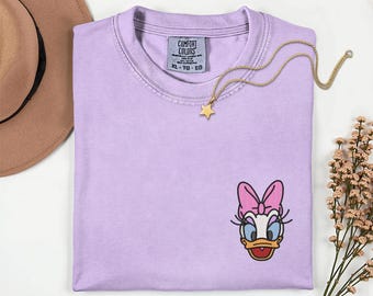 May include: A light purple t-shirt with an embroidered Daisy Duck design. The shirt has a crew neck and a small tag on the collar. A gold necklace with a star pendant is draped across the shirt.