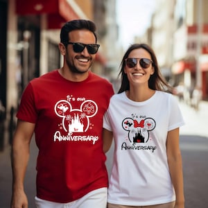 May include: A couple wearing matching t-shirts with a Disney-themed design. The red shirt says "It's Our 1st Anniversary" with a Mickey Mouse head design. The white shirt has a similar design with a Minnie Mouse bow.