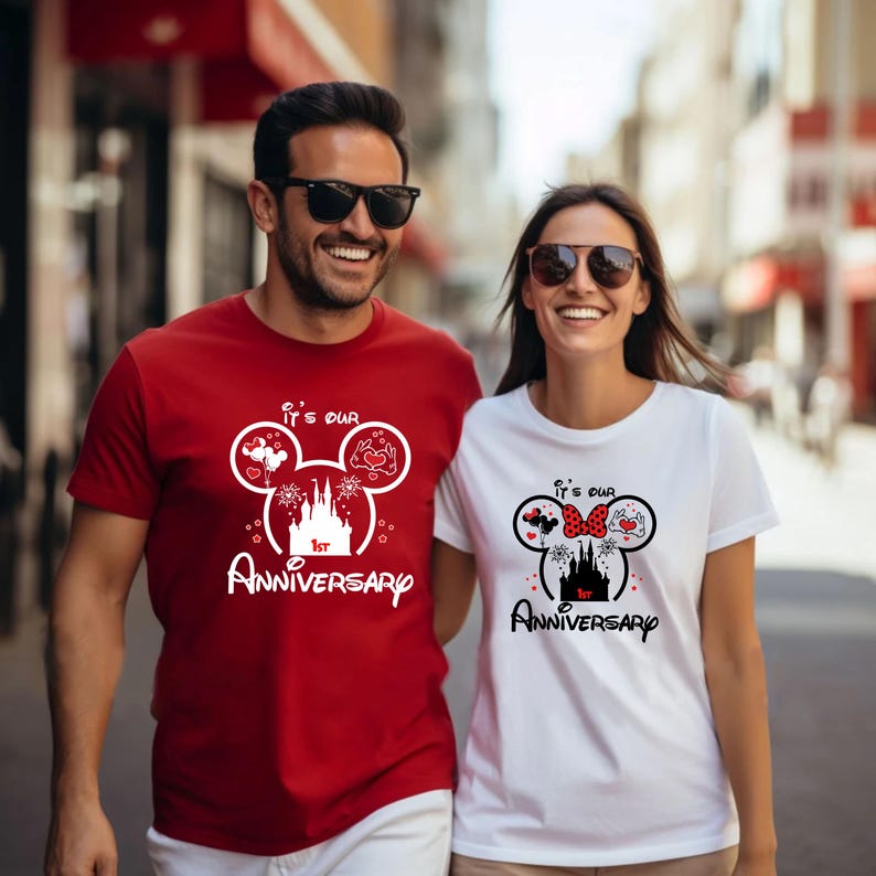 May include: A couple wearing matching t-shirts with a Disney-themed design. The red shirt says "It's Our 1st Anniversary" with a Mickey Mouse head design. The white shirt has a similar design with a Minnie Mouse bow.
