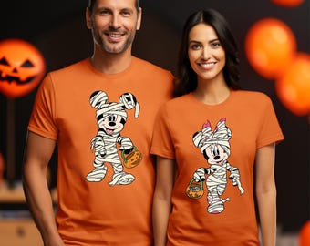 May include: Two orange short-sleeve t-shirts featuring graphic prints of cartoon mice wrapped in white bandages like mummies. One mouse wears a bow and carries a small orange pumpkin bucket, while the other mouse also carries a pumpkin bucket.
