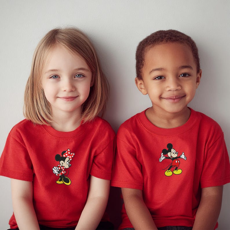 May include: Two children wearing red t-shirts featuring Disney characters. One shirt displays Minnie Mouse with a polka dot bow, and the other shows Mickey Mouse. The shirts are short-sleeved and have a classic cartoon design.