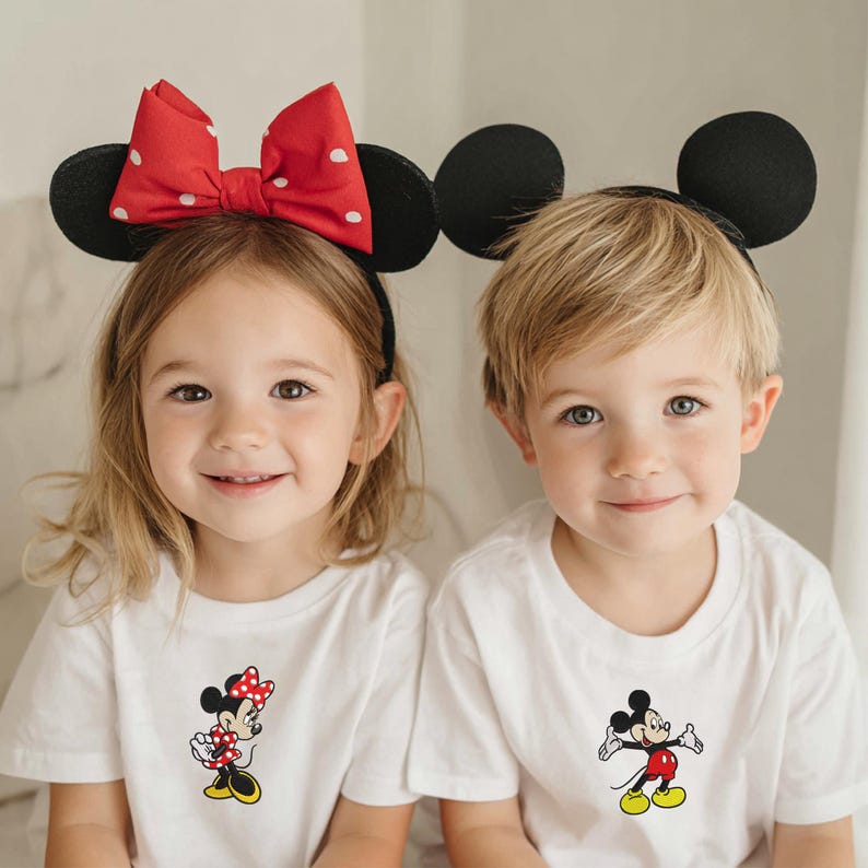 May include: Two children wearing white t-shirts with Disney character graphics. One child wears a Minnie Mouse headband with a red bow and white polka dots. The other child wears a Mickey Mouse headband with black ears.