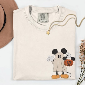 Pu&ograve; includere: T-shirt color crema con un personaggio fantasma dei cartoni animati che tiene in mano una lanterna a forma di zucca. La maglietta ha uno scollo tondo e un'etichetta con la scritta "COMFORT COLORS". Una collana con pendente a stella dorata e un cappello marrone sono anch'essi nell'immagine.