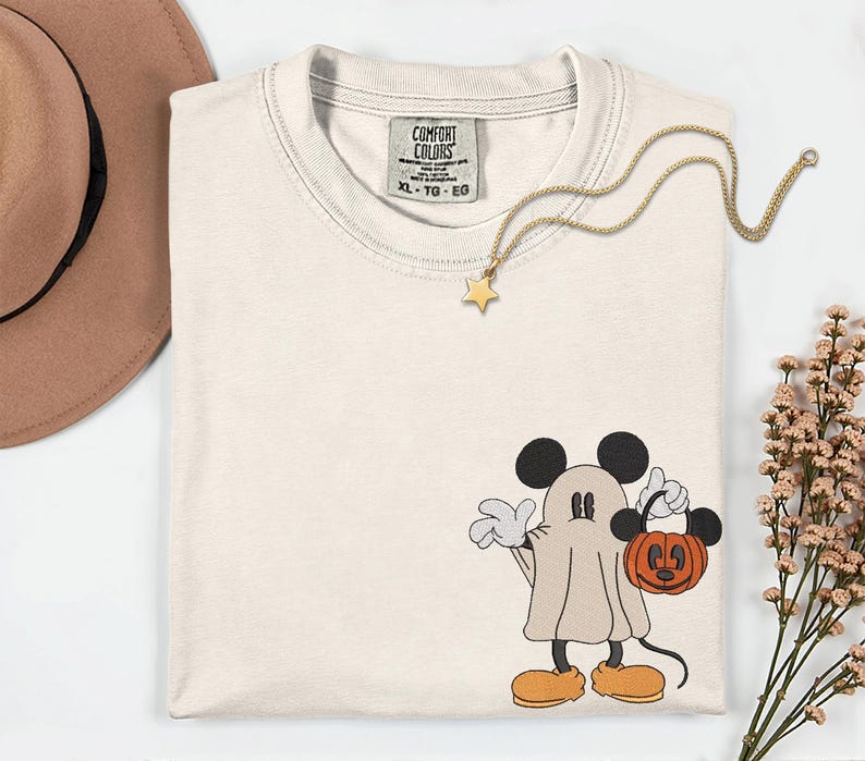 Pu&ograve; includere: T-shirt color crema con un personaggio fantasma dei cartoni animati che tiene in mano una lanterna a forma di zucca. La maglietta ha uno scollo tondo e un'etichetta con la scritta "COMFORT COLORS". Una collana con pendente a stella dorata e un cappello marrone sono anch'essi nell'immagine.