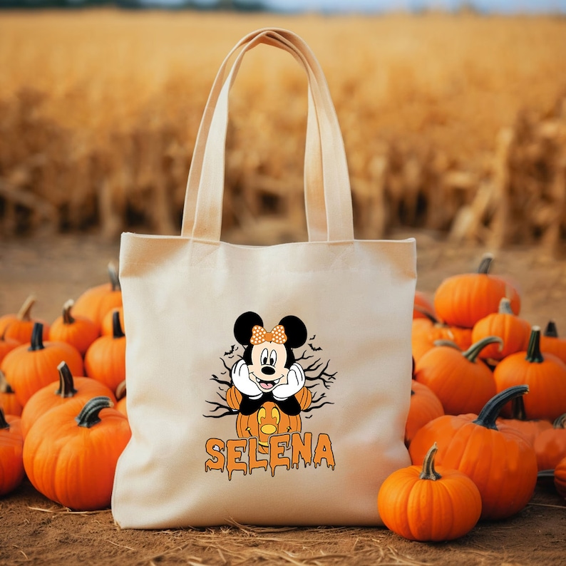 May include: A white canvas tote bag with a Halloween-themed design featuring Mickey Mouse dressed as a pumpkin. The bag has the name "SELENA" printed in orange dripping letters below the image.