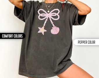 May include: A charcoal gray short-sleeve t-shirt featuring a graphic print of a large bow made of pink pearls. Two strands of pearls hang from the bow, one ending in a multi-colored starfish and the other in a pink seashell. The text reads COMFORT COLORS and PEPPER COLOR.