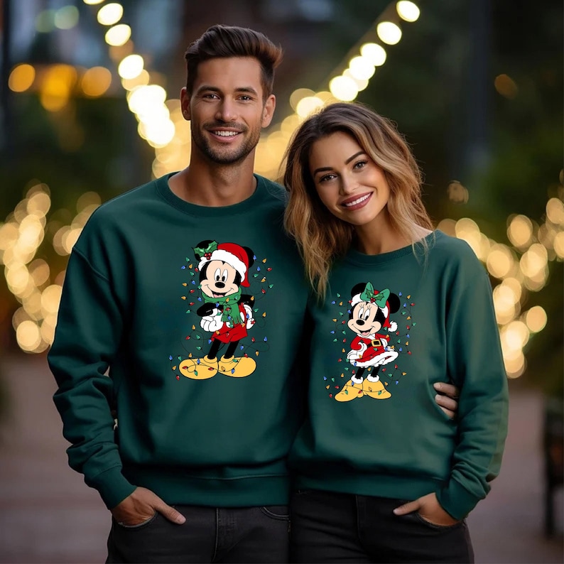 K&ouml;nnte beinhalten: Zwei Personen tragen passende dunkelgr&uuml;ne Sweatshirts. Jedes Sweatshirt zeigt ein Cartoon-Design von Mickey und Minnie Mouse in Weihnachtskleidung, umgeben von bunten Lichterketten. Der Hintergrund ist mit Bokeh-Lichtern verschwommen.