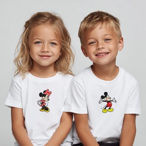May include: Two children wearing white t-shirts with cartoon character designs. One shirt features Minnie Mouse in a red polka-dot bow and dress. The other shirt displays Mickey Mouse in red shorts and yellow shoes. Both shirts are short-sleeved.