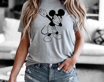May include: A gray t-shirt featuring a black and white graphic of Mickey Mouse doing the dab. The cartoon character is wearing white gloves, shorts, and shoes. The shirt is paired with denim shorts.
