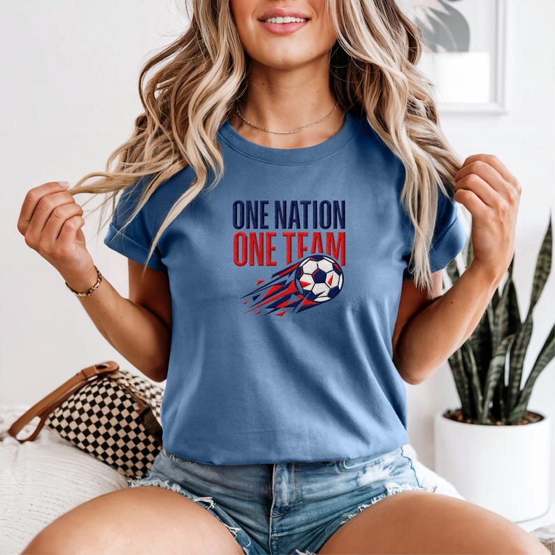 May include: A light blue t-shirt with the text "ONE NATION ONE TEAM" in red and blue. A soccer ball with red and blue streaks is below the text. The shirt is a crew neck style.