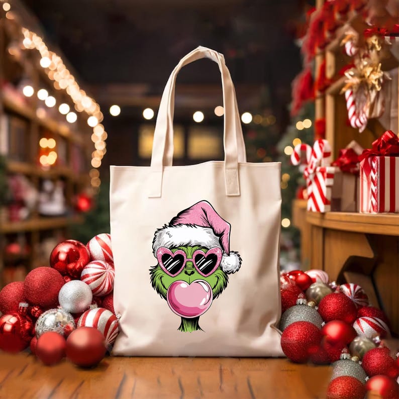 May include: A cream-colored tote bag featuring a cartoon Grinch face wearing a pink Santa hat and heart-shaped sunglasses. The Grinch is blowing a pink bubblegum bubble. Christmas ornaments surround the bag.