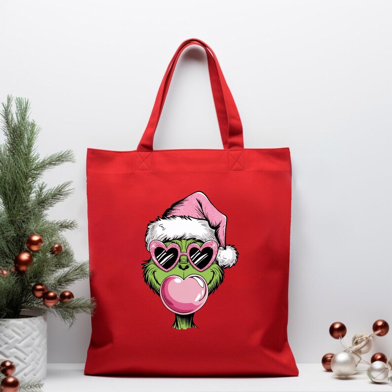 May include: Red tote bag featuring a cartoon Grinch character wearing a pink Santa hat and heart-shaped sunglasses, blowing a bubblegum bubble. The bag has a simple design with a solid red color and sturdy handles.