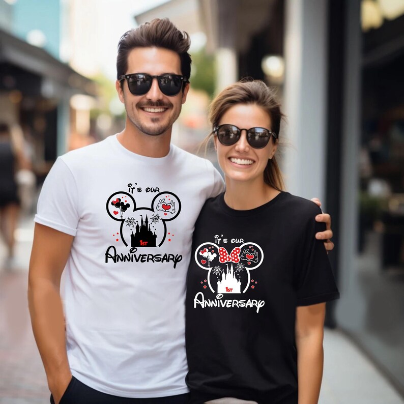May include: A couple wearing matching white and black t-shirts with a Mickey Mouse-inspired design. The shirts feature a castle, hearts, balloons, and the text "It's our 1st Anniversary."