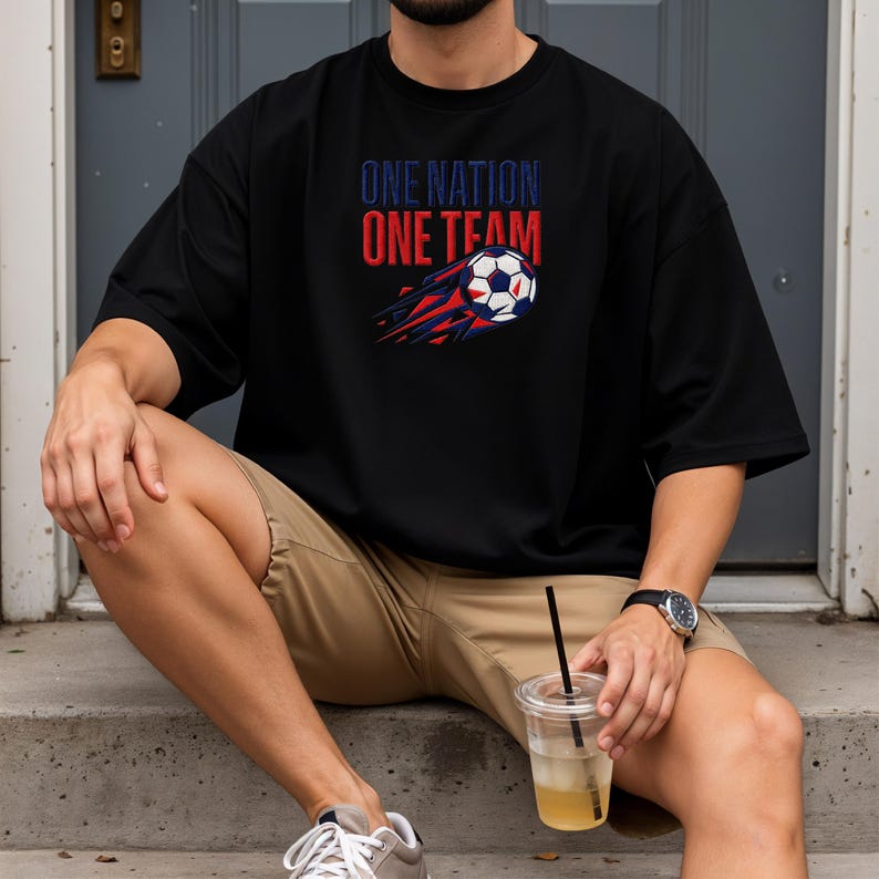 May include: Black t-shirt with the text "ONE NATION ONE TEAM" in red and blue. A soccer ball with red and blue streaks is featured below the text. The shirt is a casual style.