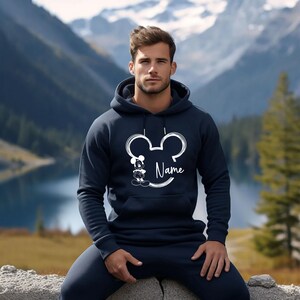 K&ouml;nnte beinhalten: Ein marineblauer Hoodie mit einer wei&szlig;en Umrandung von Mickey Mouses Kopf und Ohren. Minnie Mouse steht innerhalb der Umrandung. Der Text "Name" steht unterhalb der Umrandung.