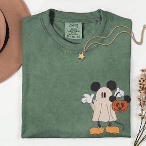 Pu&ograve; includere: T-shirt verde Comfort Colors con grafica di Mickey Mouse a tema Halloween. Mickey &egrave; raffigurato come un fantasma, con in mano un sacchetto di dolcetti a forma di zucca. Nel'immagine sono presenti anche una collana a stella dorata e un cappello marrone.