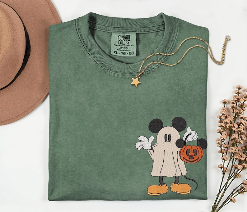 Pu&ograve; includere: T-shirt verde Comfort Colors con grafica di Mickey Mouse a tema Halloween. Mickey &egrave; raffigurato come un fantasma, con in mano un sacchetto di dolcetti a forma di zucca. Nel'immagine sono presenti anche una collana a stella dorata e un cappello marrone.