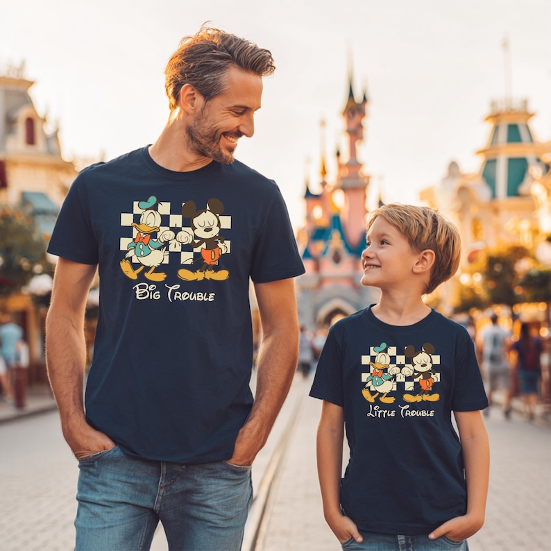 May include: Navy blue t-shirts with a cartoon graphic of Donald Duck and Mickey Mouse on a checkered background. The shirt on the left says "Big Trouble" and the shirt on the right says "Little Trouble".