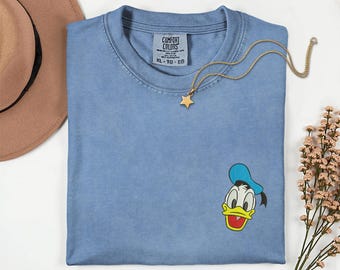 May include: A light blue Comfort Colors t-shirt with a small embroidered Donald Duck graphic. The shirt has a crew neck and short sleeves. A gold star pendant necklace is draped across the shirt. The tag reads "Comfort Colors XL-TG-EG".