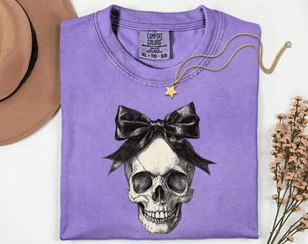 May include: A purple short-sleeved t-shirt featuring a graphic print of a human skull adorned with a large black bow. The shirt has a Comfort Colors brand label on the inside neck area, indicating size XL. A gold-toned star pendant necklace is placed on the shirt.