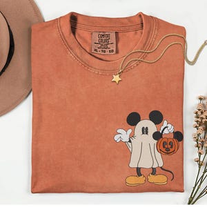 Pu&ograve; includere: T-shirt Comfort Colors color ruggine con un fantasma di Topolino che tiene in mano una lanterna a forma di zucca. Una collana con pendente a stella dorata &egrave; appoggiata sulla maglietta. La maglietta ha un'etichetta con la scritta "Comfort Colors". Un cappello marrone &egrave; nell'angolo in alto a sinistra.