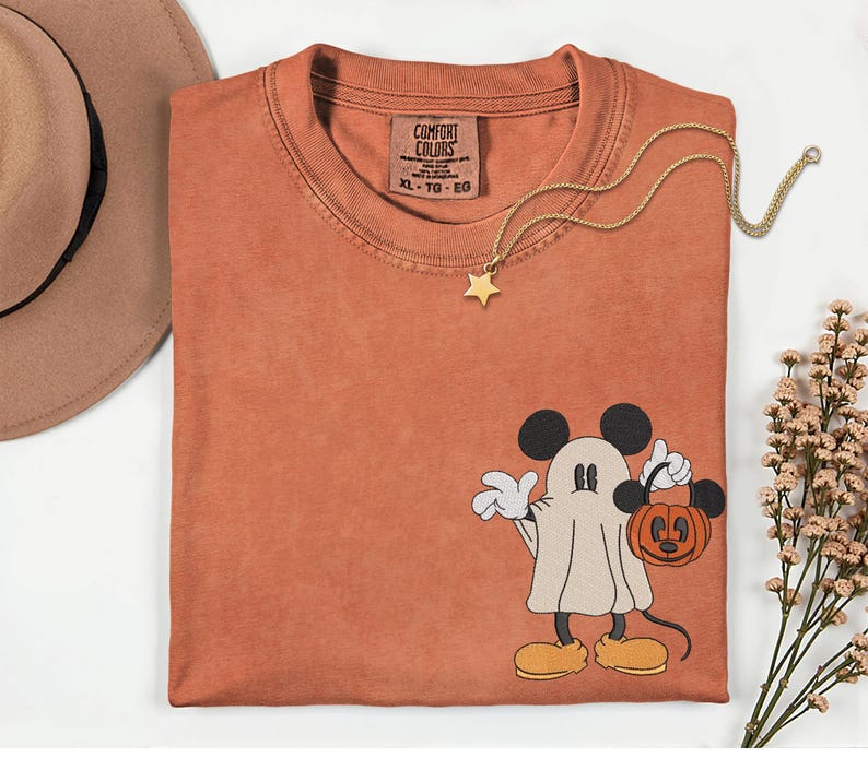 Pu&ograve; includere: T-shirt Comfort Colors color ruggine con un fantasma di Topolino che tiene in mano una lanterna a forma di zucca. Una collana con pendente a stella dorata &egrave; appoggiata sulla maglietta. La maglietta ha un'etichetta con la scritta "Comfort Colors". Un cappello marrone &egrave; nell'angolo in alto a sinistra.