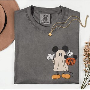 Pu&ograve; includere: T-shirt grigio Comfort Colors con un design di Mickey Mouse a tema Halloween. Mickey &egrave; vestito da fantasma e tiene in mano una zucca arancione. Una collana con pendente a stella dorata &egrave; drappeggiata sulla maglietta.