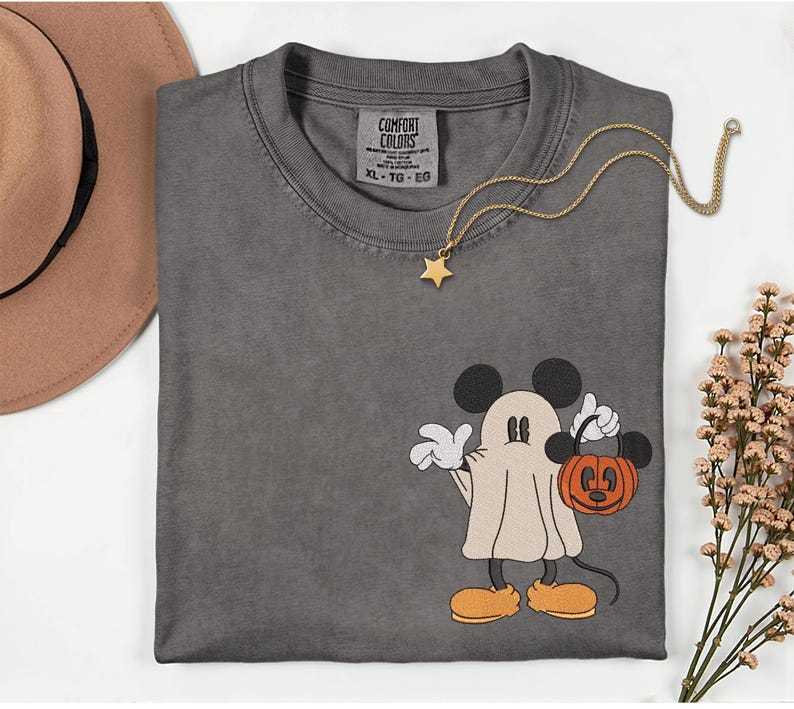 Pu&ograve; includere: T-shirt grigio Comfort Colors con un design di Mickey Mouse a tema Halloween. Mickey &egrave; vestito da fantasma e tiene in mano una zucca arancione. Una collana con pendente a stella dorata &egrave; drappeggiata sulla maglietta.