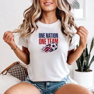 May include: White t-shirt with the text "ONE NATION ONE TEAM" in red and blue. A soccer ball with red and blue streaks is below the text. The shirt is a crew neck style.