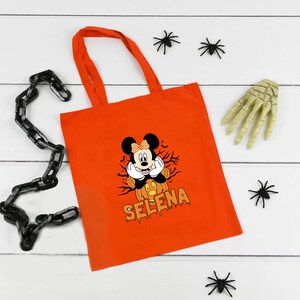 May include: Orange tote bag with a black and white image of Mickey Mouse dressed as a pumpkin. The text "Selena" is printed in black below the image.
