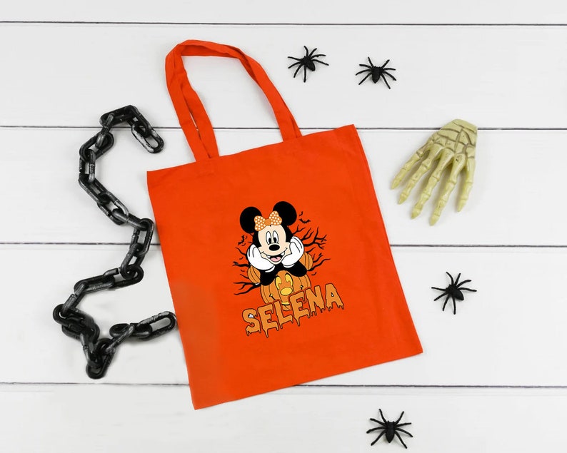 May include: Orange tote bag with a black and white image of Mickey Mouse dressed as a pumpkin. The text "Selena" is printed in black below the image.