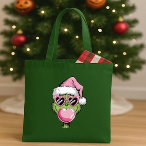 May include: A green tote bag featuring a cartoon character with a pink Santa hat, heart-shaped sunglasses, and a pink bubblegum bubble. The bag has a gift wrapped in red and green plaid inside.