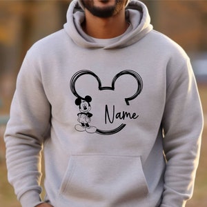 K&ouml;nnte beinhalten: Ein grauer Hoodie mit einer schwarzen Umrandung von Mickey Mouses Kopf und Ohren. Das Wort "Name" steht innerhalb der Ohren. Ein kleines Bild von Mickey Mouse befindet sich links neben der Umrandung.
