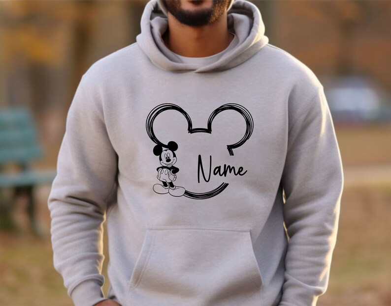 K&ouml;nnte beinhalten: Ein grauer Hoodie mit einer schwarzen Umrandung von Mickey Mouses Kopf und Ohren. Das Wort "Name" steht innerhalb der Ohren. Ein kleines Bild von Mickey Mouse befindet sich links neben der Umrandung.