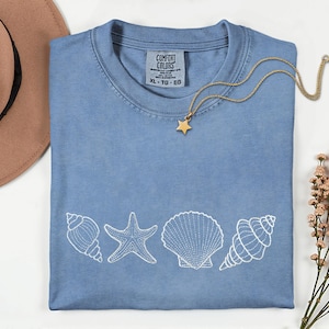 May include: A light blue t-shirt with a white seashell design and a gold star pendant necklace. The shirt has the text "COMFORT COLORS" on the tag. The design includes a starfish and various seashells.