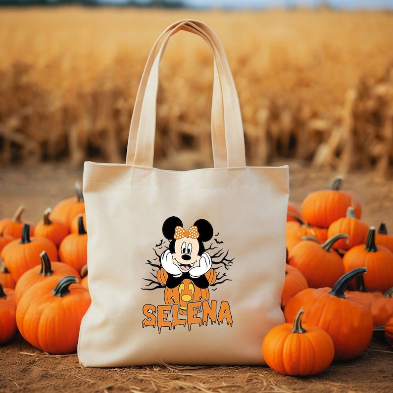 May include: A white canvas tote bag with a Mickey Mouse character dressed as a pumpkin. The bag has the name "SELENA" written in orange dripping letters. The bag is surrounded by orange pumpkins.