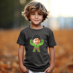 K&ouml;nnte beinhalten: Ein dunkelgraues T-Shirt mit einem Cartoon-Bild eines gr&uuml;nen Hulks mit dem Gesicht eines Jungen. Der Hulk zeigt seine Muskeln und tr&auml;gt einen braunen Lendenschurz.