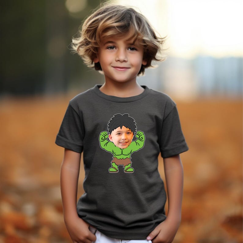 K&ouml;nnte beinhalten: Ein dunkelgraues T-Shirt mit einem Cartoon-Bild eines gr&uuml;nen Hulks mit dem Gesicht eines Jungen. Der Hulk zeigt seine Muskeln und tr&auml;gt einen braunen Lendenschurz.