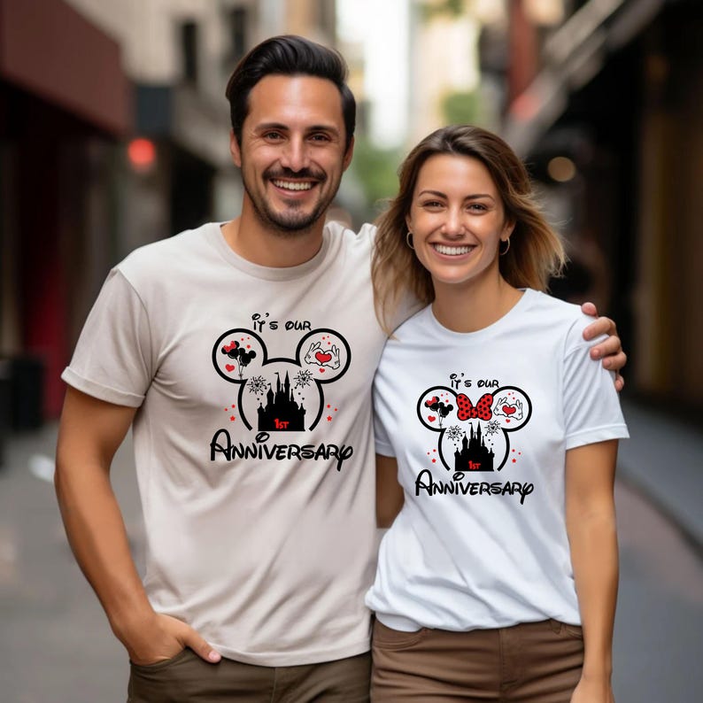 May include: Matching beige and white t-shirts with a Mickey Mouse-inspired design for a 1st anniversary. The design includes a castle, hearts, and the text "It's our 1st Anniversary" in black and red.
