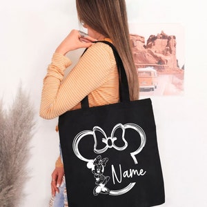 May include: Black tote bag with a white outline of Minnie Mouse's head and bow. The bag has the word "Name" written below the image.